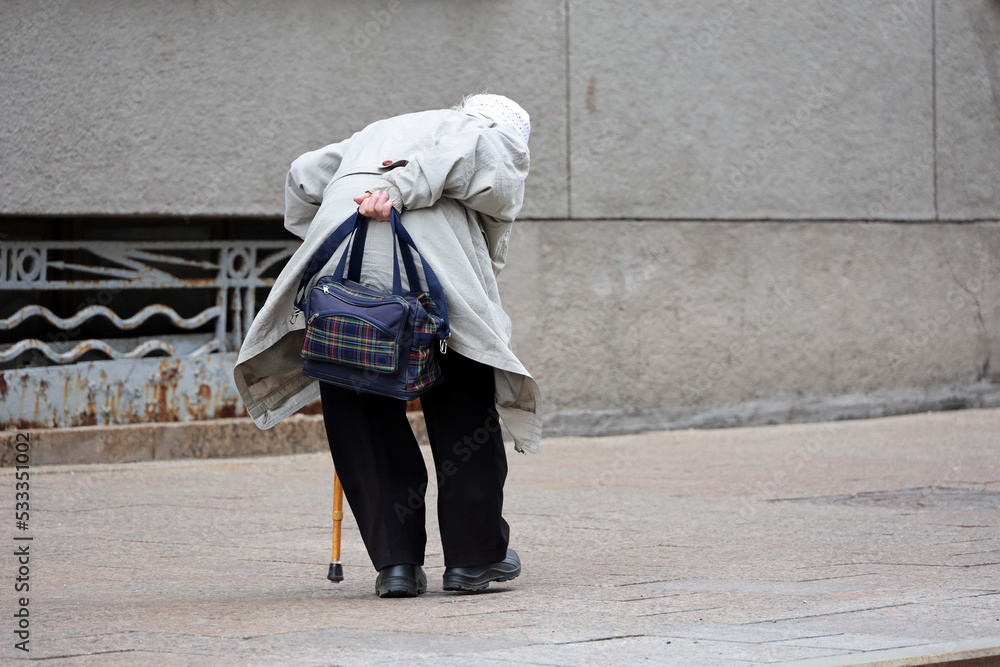 Elderly woman walking bent low with a cane and bag on a city street. Life of old people in