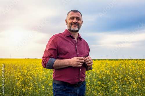 Middle age farmer standing in rapeseed field examining crop.