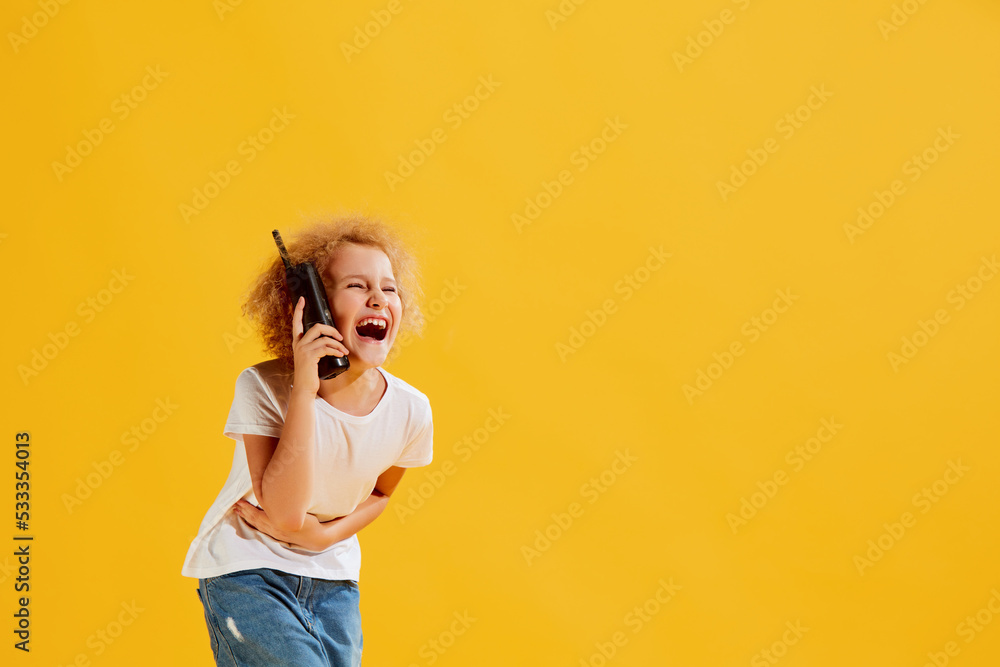 Laughing. Cute little girl, kid using retro mobile phone isolated over yellow background. Concept of children emotions, fashion
