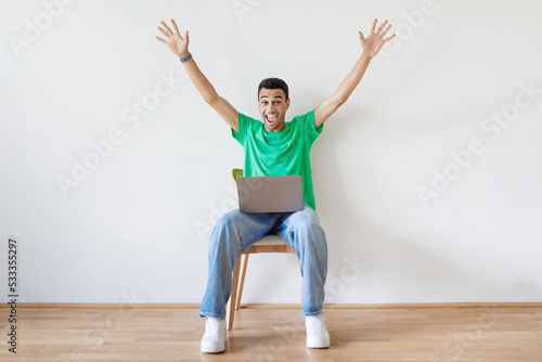 Fototapeta Naklejka Na Ścianę i Meble -  Emotional arab man sitting on chair with laptop and raising hands up, happy guy got dream job or great news