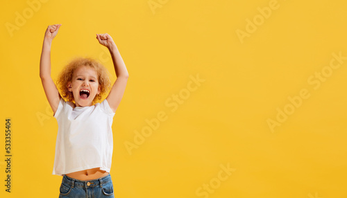 Tablou canvas Thrilled little girl, kid shouting in rejoice and raising hands up isolated over yellow background
