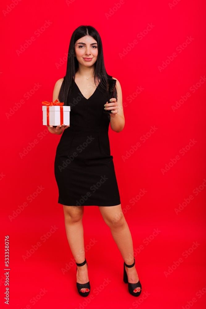 Congratulations to colleagues on Christmas. A young woman in a dress and heels with gifts and a glass of champagne on a red background. Congratulations to colleagues on Christmas. A young woman in a dress and heels with gifts and a glass of champagne on a red background.