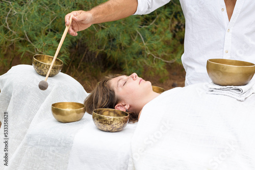 Young woman laying on a massage bed with tibetan singing bowl outdoor. A master of sound massage therapy doing a Buddhist healing practices, male hand holding a felt clapper