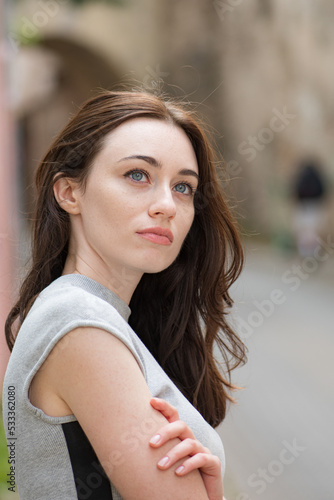 Beautiful Ukrainian girl with red hair, blue eyes and freckles in a short grey summer dress in the city old town with old houses on background, vertical