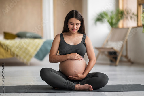 Maternity Concept. Young Pregnant Female Sitting On Yoga Mat And Embracing Belly