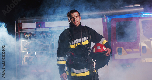Εκτύπωση καμβά Middle plan of tired Caucasian young man fire fighter taking off helmet and walking out from fire at the red truck and looking into camera