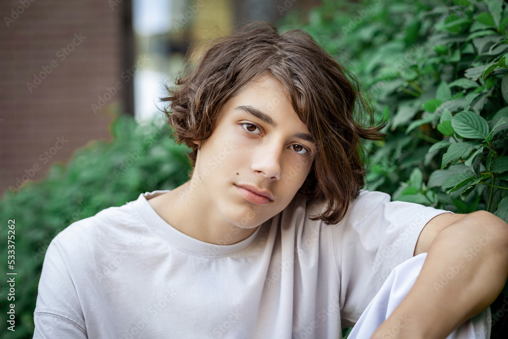 Portrait of a teenager in a beige T-shirt against a background of green foliage. A young guy with long hair on a summer city street. Portrait of a teenager in a beige T-shirt against a background of green foliage. A young guy with long hair on a summer city street.