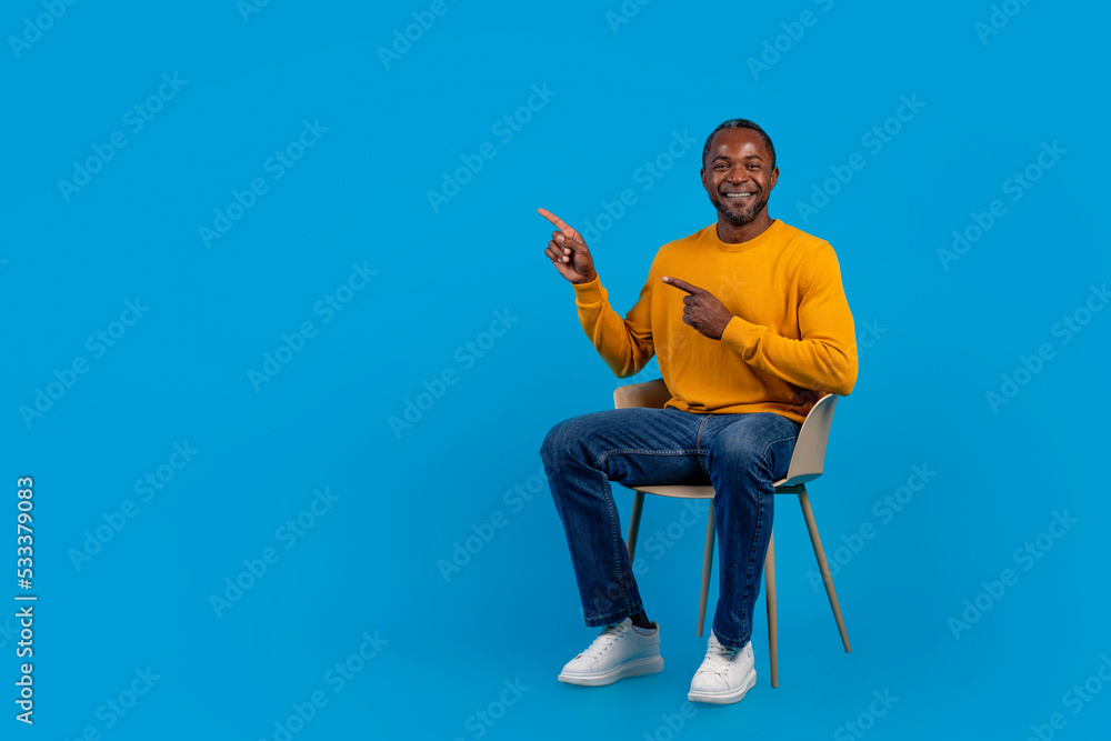 Positive black man sitting on chair, pointing at copy space Positive black man sitting on chair, pointing at copy space