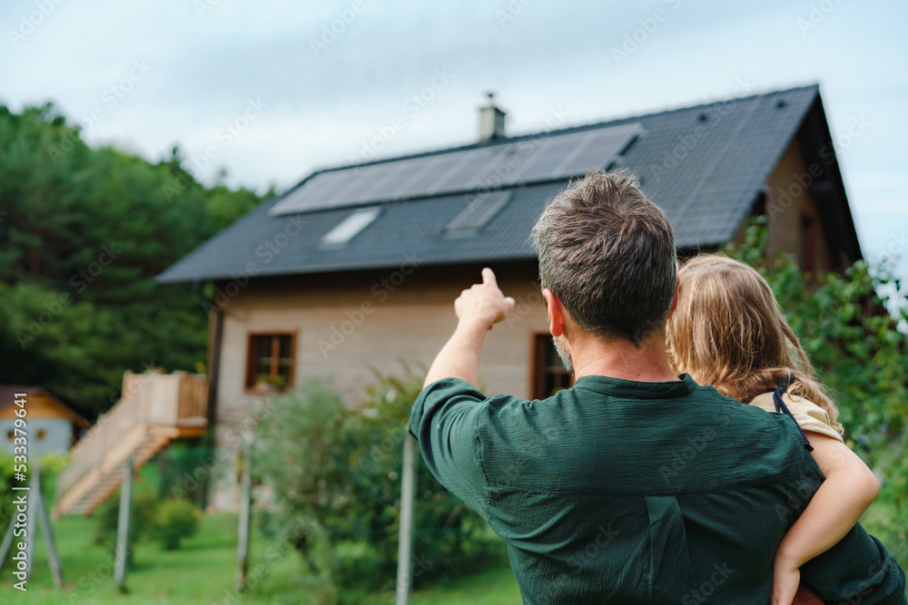 Rear view of dad holding her little girl in arms and showing at their house with installed solar panels. Alternative energy, saving resources and sustainable lifestyle concept. - obrazy, fototapety, plakaty 