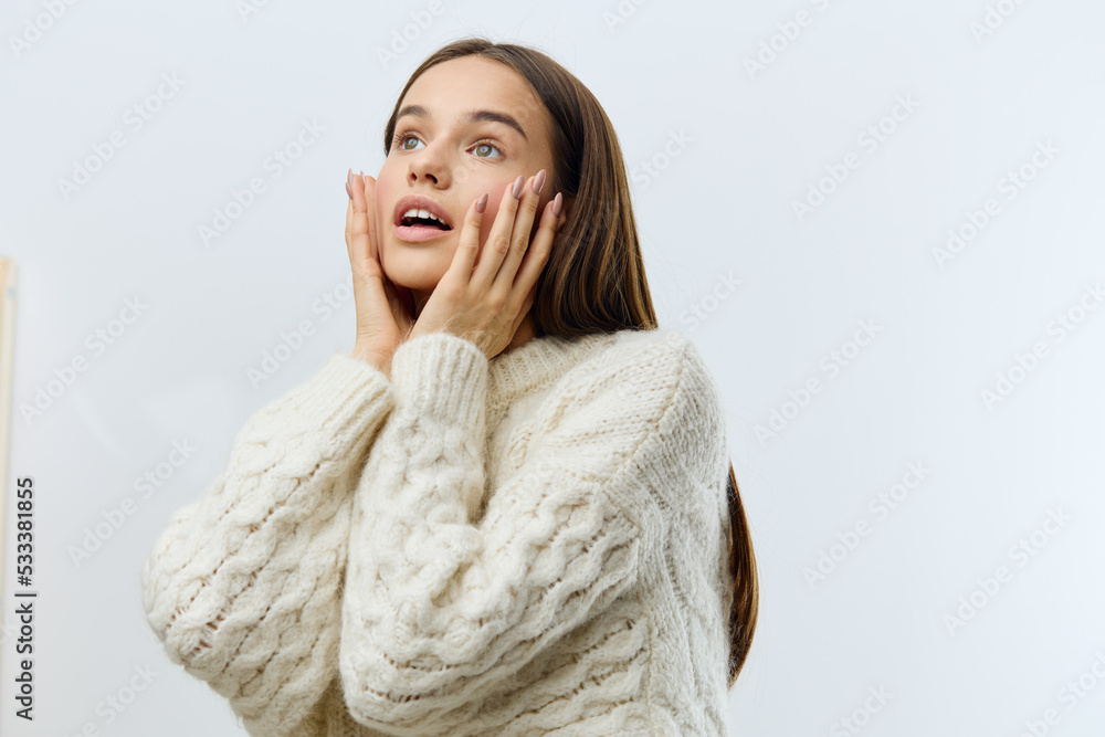 a beautiful, attractive, emotional woman stands on a light background in a white sweater and looks admiringly to the side touching her face with her hands a beautiful, attractive, emotional woman stands on a light background in a white sweater and looks admiringly to the side touching her face with her hands