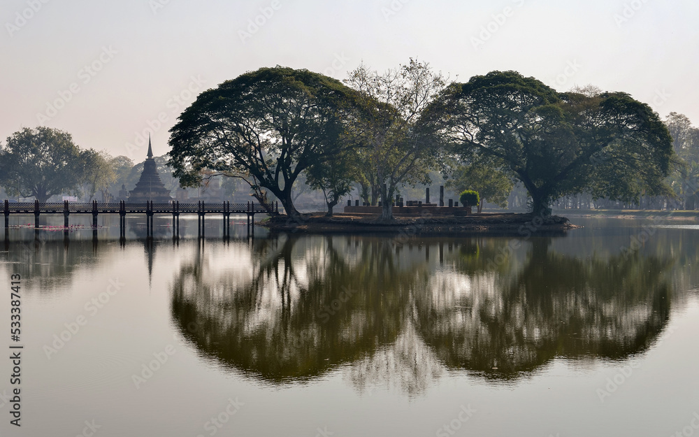 Beautiful morning view of Wat Traphang Ngoen temple at sunrise in Sukhothai Historical Park, Thailand. Old trees reflecting on water like a mirror.