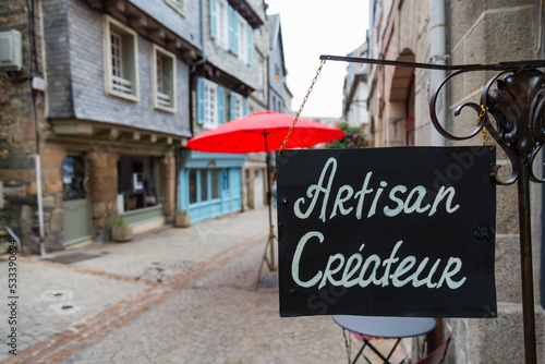 Shop sign outside a traditional shop in Morlaix, Brittany, France. Text translation "artisan creator". Ancient street of the historical centre of the village in the background.