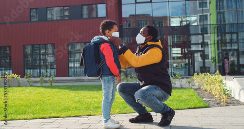 Young man dad in mask standing outdoor at schoolyard and putting on cute little boy mask before lessons. Father and junior student son looking at camera and smiling. Quarantine education concept