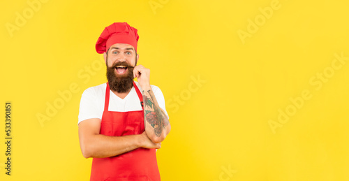 Happy man in red toque and apron twirling moustache yellow background copy space, cook
