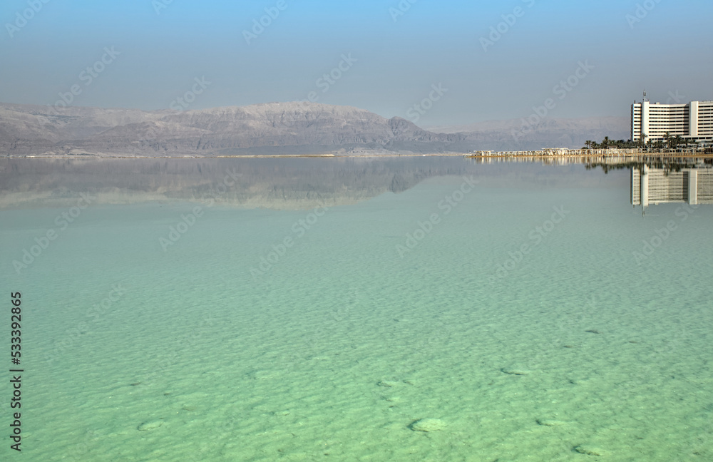 Wide view on the Dead Sea, Israel. Turquoise water of a salt lake, white gazebos on a sandy beach and a hotel against a background of mountain range reflected on the salty water