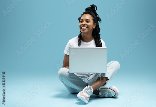 Quadro su tela Happy young woman sitting on the floor with crossed legs and using laptop on blue background