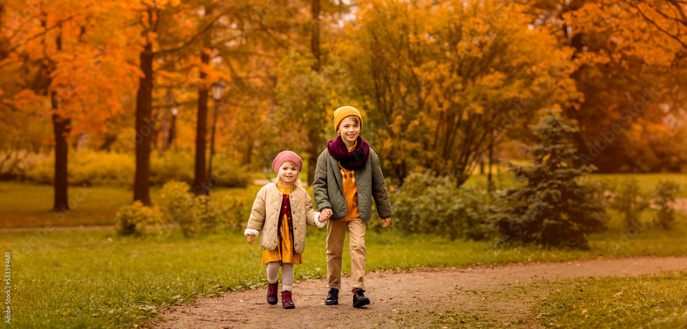 Fototapeta premium Autumn park children boy and girl walking in fall golden leaves path October season. Kids wearing knitted casual stylish clothes. Positive emotion. Copy space