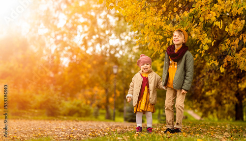 Autumn park children boy and girl stay on path, laugh and looking at camera in fall golden leaves meadow October season. Kids wearing knitted casual stylish clothes. Positive emotion. Copy space