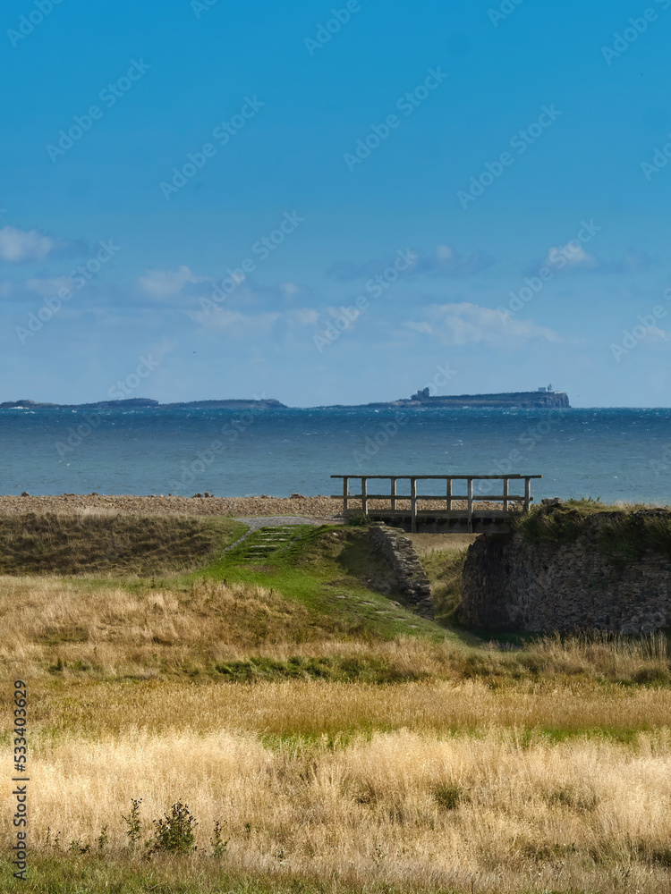 The legendary landscape of Northumberland, a bridge across Lindisfarne's beautifully stark heathland, the cold blue of the North Sea and the rugged silhouette of the Farne Islands on the horizon.