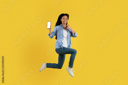 Cheerful young japanese lady student in casual jumping, pointing finger at smartphone with empty screen photo