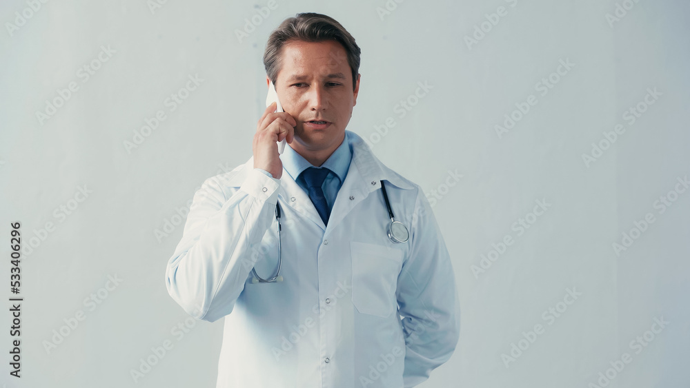doctor in white coat standing with hand behind back while talking on smartphone on grey. doctor in white coat standing with hand behind back while talking on smartphone on grey.