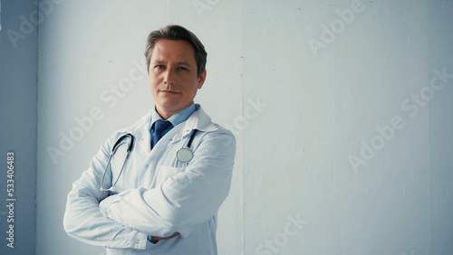 confident doctor in white coat standing with crossed arms near grey wall.