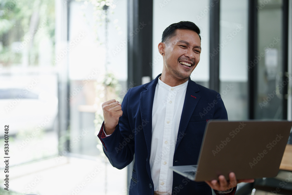 Portrait of an Asian male business owner standing with a computer showing happiness after a successful investment Portrait of an Asian male business owner standing with a computer showing happiness after a successful investment
