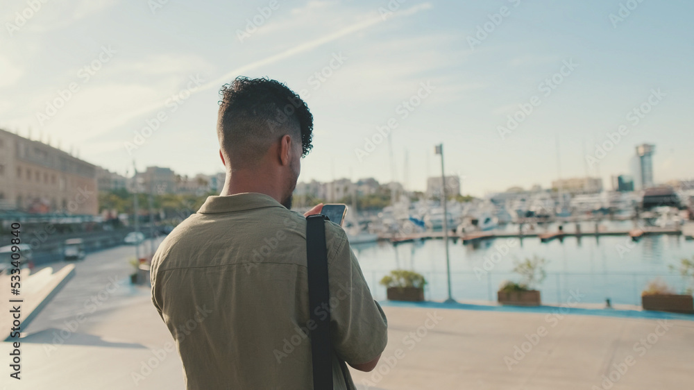 Close up, young man dressed in an olive-colored shirt is standing in the port looking at the yachts and shooting on smartphone Close up, young man dressed in an olive-colored shirt is standing in the port looking at the yachts and shooting on smartphone