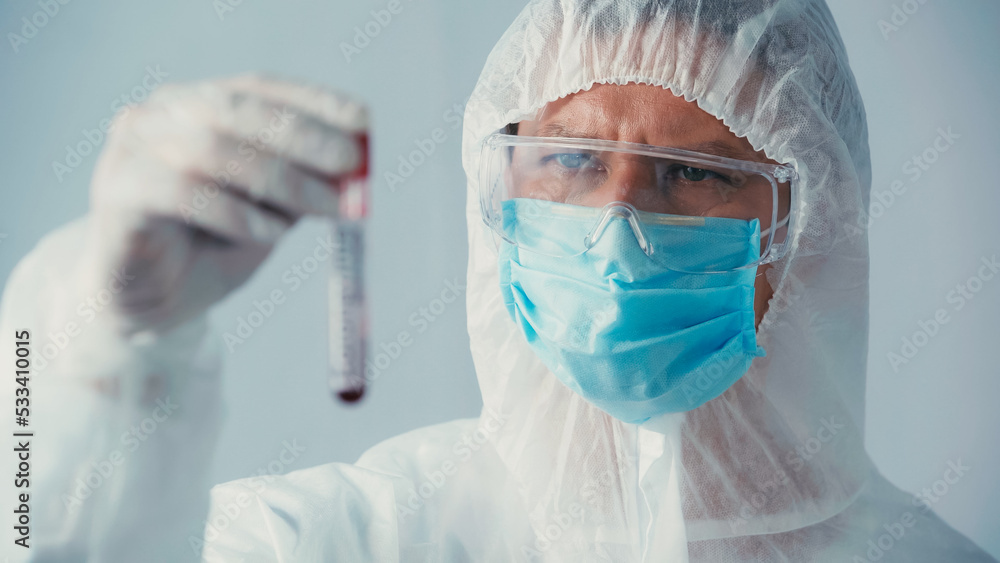 virologist in hazmat suit and goggles looking at blurred test tube isolated on grey. virologist in hazmat suit and goggles looking at blurred test tube isolated on grey.