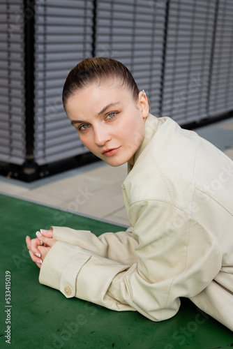 Young woman in trench coat looking at camera near ping-pong table outdoors.