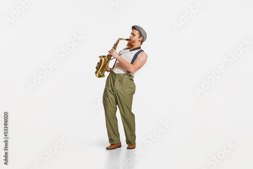 Young musician in retro style outfit playing saxophone isolated over white studio background. Concept of music, hobby, festival.