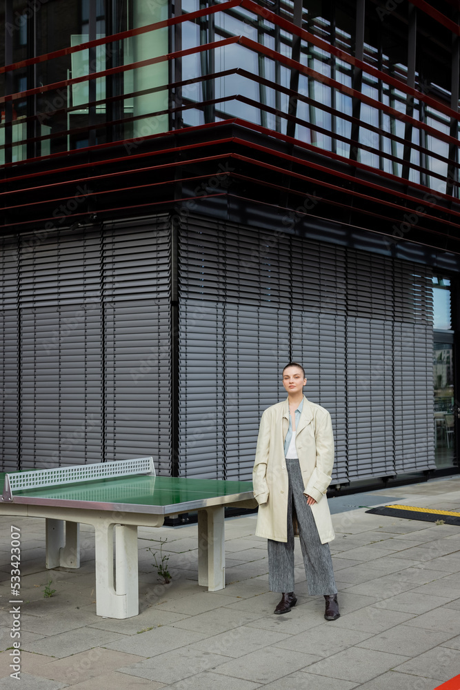 Stylish woman in trench coat standing near ping-pong table outdoors in Berlin.