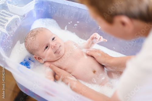 Baby bath time. Close-up detail view of mother bathing cute little peaceful baby in tub with water and bubbles lather.