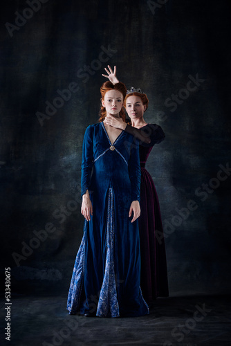 Portrait of two beautiful women in image of queens isolated over dark background. Envy  rivalry  intrigue
