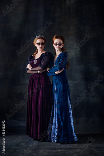 Portrait of two beautiful women in image of queen and princess in stylish sunglasses isolated over dark background