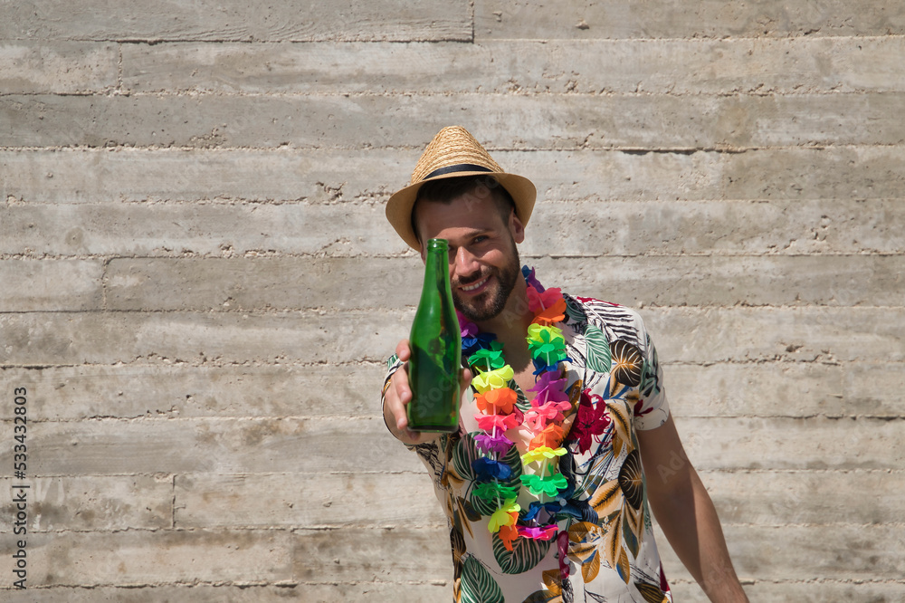 Young and handsome man, blue eyes, with beard, hat, Hawaiian shirt and flower necklace and offering a bottle of beer on a gray background. Concept vacation, party, travel, drink. Selective focus.