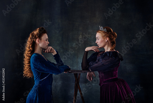 Portrait of two beautiful women in image of queens isolated over dark background. Confrontation, eye to eye photo