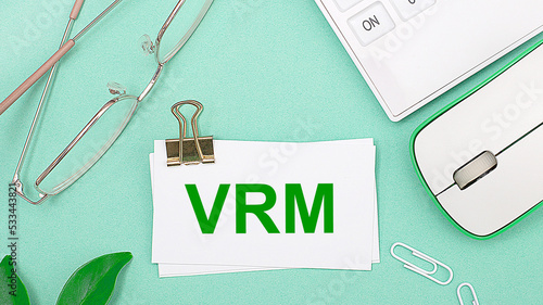Photos On a light green background there is a white calculator, a computer mouse, green leaves of a plant, gold-rimmed glasses and a white card with text VRM Vendor Relationship Management