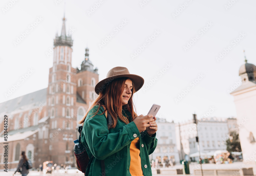 Beautiful stylish woman in hat walking on Market Square in Krakow on autumn day and holding mobile phone. Phone Communication. Urban lifestyle concept. Check social networks, send sms.