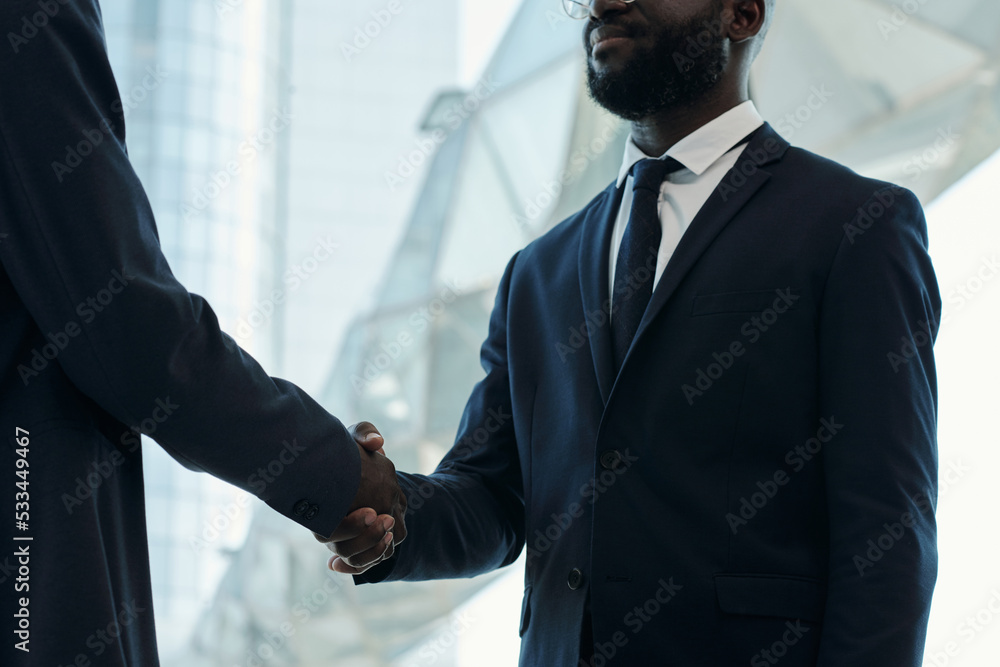 Cropped shot of young African American businessman in dark blue suit handshaking with new business partner after negotiating Cropped shot of young African American businessman in dark blue suit handshaking with new business partner after negotiating