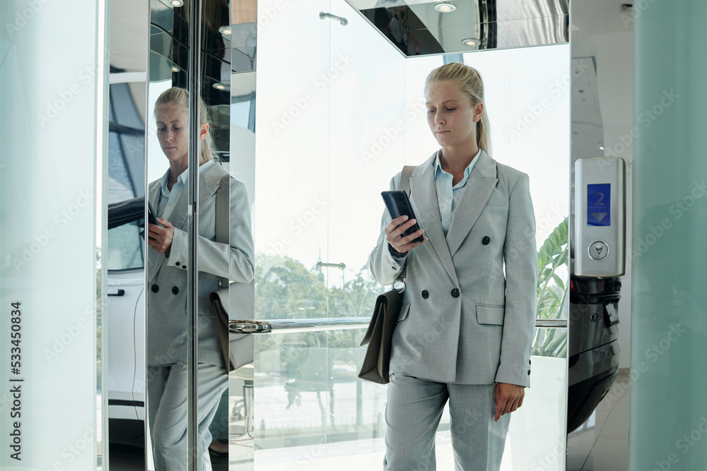 Young modern businesswoman in grey formalwear texting in mobile phone while going out of elevator on second floor of office center Young modern businesswoman in grey formalwear texting in mobile phone while going out of elevator on second floor of office center