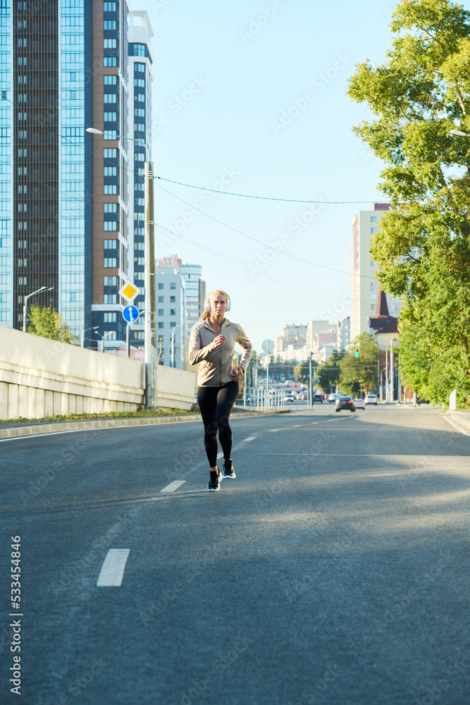 Young sportswoman in activewear running along asphalt road in the morning against modern buildings and blue sky Young sportswoman in activewear running along asphalt road in the morning against modern buildings and blue sky