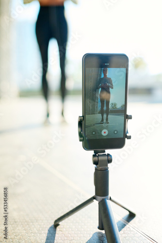 Tripod with smartphone during video record of sportswoman having outdoor workout while standing in urban environment