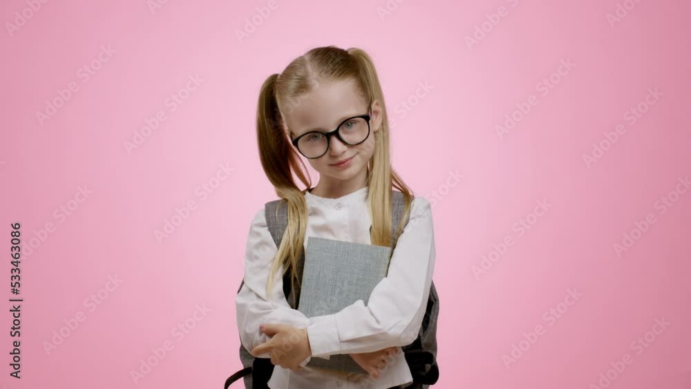 Little genius. Cute pretty little schoolgirl carrying backpack and books, fixing her eyeglasses and smiling to camera Little genius. Cute pretty little schoolgirl carrying backpack and books, fixing her eyeglasses and smiling to camera