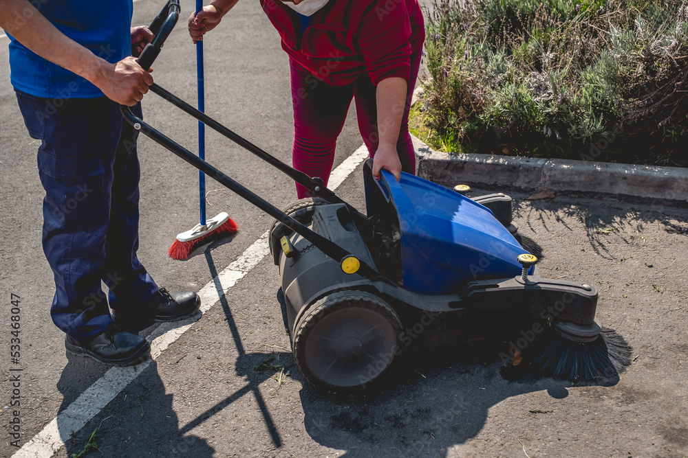 Two young latino janitorial workers (man and woman) in uniform cleaning in parking lot with outdoor industrial vacuum cleaner