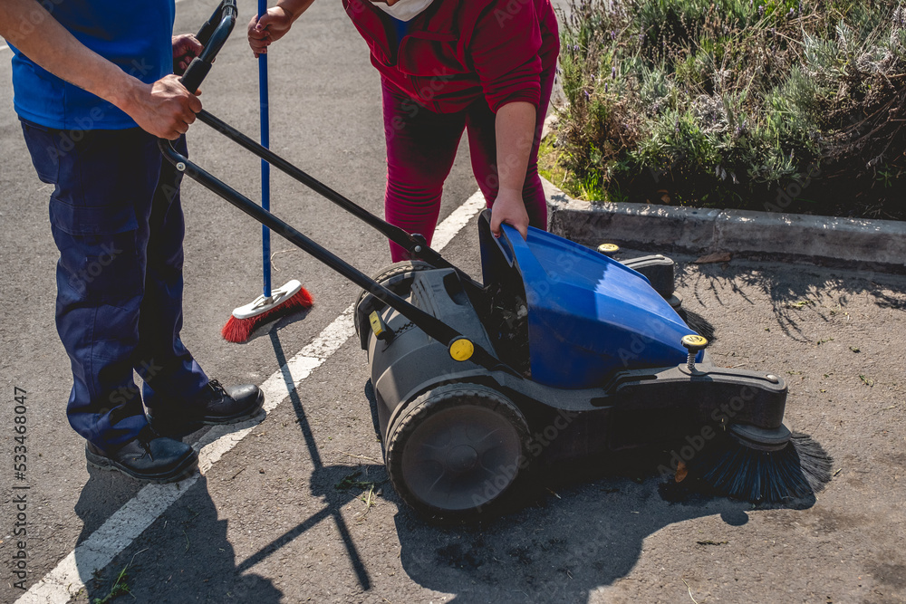 Obraz premium Two young latino janitorial workers (man and woman) in uniform cleaning in parking lot with outdoor industrial vacuum cleaner