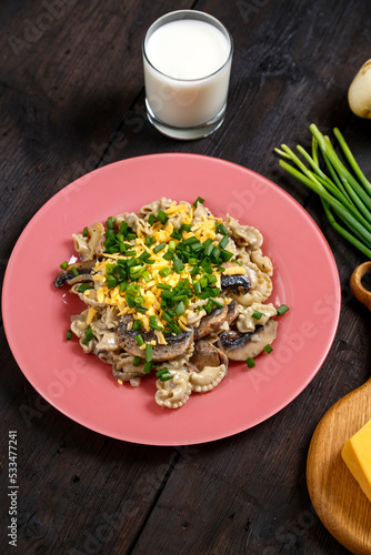 Pasta in a creamy sauce with mushrooms in a plate on the table next to a glass of cream, green onions and champignons.