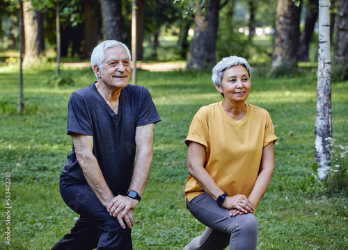 Senior spouses doing sportive exercises outdoor in summer park in the morning. Hoary healthy older couple warming-up, doing squats smile enjoy active lifestyle outside