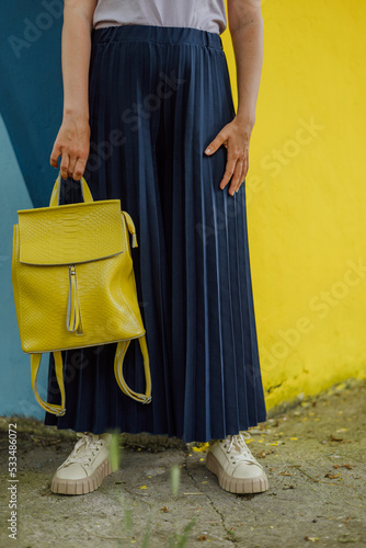 vertical portrait of a young woman with a yellow backpack in the summer on the street against the background of a colored wall in blue and yellow