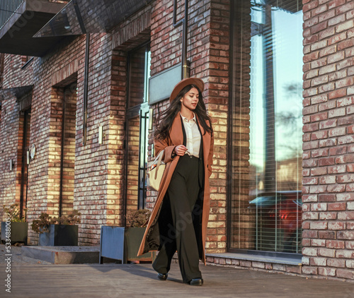 Asian business woman in long beige coat and hat walking down street in loft style, on background of brick wall, classic retro style © okostia